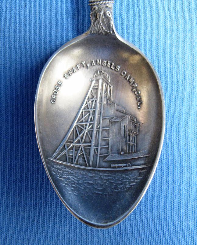 Souvenir Mining Spoon Bowl Cross Shaft.JPG - SOUVENIR MINING SPOON CROSS SHAFT UTICA MINE - Sterling silver spoon, 5 1/8 in. long, embossed mining scene in bowl with engraved CROSS SHAFT, ANGELS CAMP, CAL. in bowl, ca. 1900, back with sterling marking, handle has gold pan with crossed pick and shovel at top and a silver rope extending from a bucket ontop and wrapped around handle shaft  [The Utica Mine, one on the best-known gold mines in the Mother Lode, is located within the city of Angels Camp in southwestern Calaveras County, California. The gold mines in and around Angels Camp are part of the Angels Camp mining district, which is credited with producing at least $30 million in gold. The Utica Mine alone is thought to have produced in excess of $17 million at the old price of gold and during the 1890s, the Utica Mine was one of the most productive in the nation. The surface of the Utica claim was mined during the early part of the gold rush, but large-scale development was only conducted between 1893 and 1915 by the Utica Gold Mining Company.  In the 1880s Charles D. Lane gained control of the Utica claim and along with partners Walter Hobart and Alvinza Hayward, organized the Utica Mining Company and started large scale development in 1893. The other claims that now constitute the Utica Mine (Brown, Confidence, Dead Horse, Jackson, Little Nugget, Raspberry, Stickle, and Washington claims) were gradually acquired and the mine was developed on a major scale with more than 500 men on the payroll.  In 1896, the Cross shaft was sunk in hard rock to provide a permanent opening for the mine.  By 1900, the Cross vertical shaft bottomed at 1312 feet.  Details of mine workings are sketchy, but there are thought to be over 100 miles of underground workings.  Operations continued into late 1915 when the mine was shut down.  Except for mill cleanups and small amounts of gold recovered from the dump in the 1930s, the mine has been idle since.]